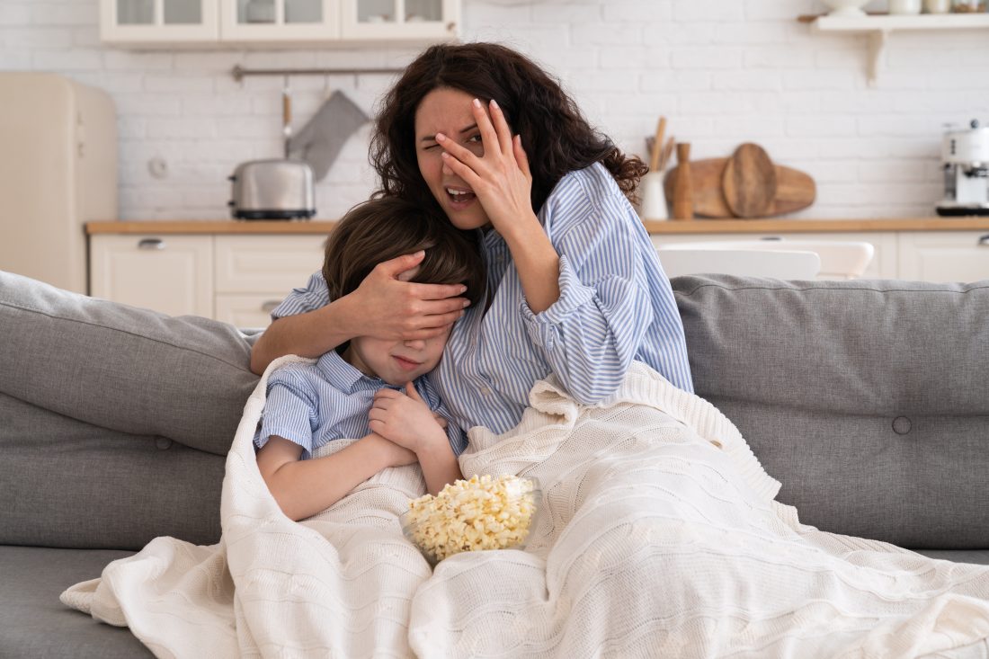 Frightened mom and little kid son with bowl of popcorn watching scary scene in movie closing their eyes, sitting on sofa at home. Mother hugs the child, watches a horror movie, feels fear.